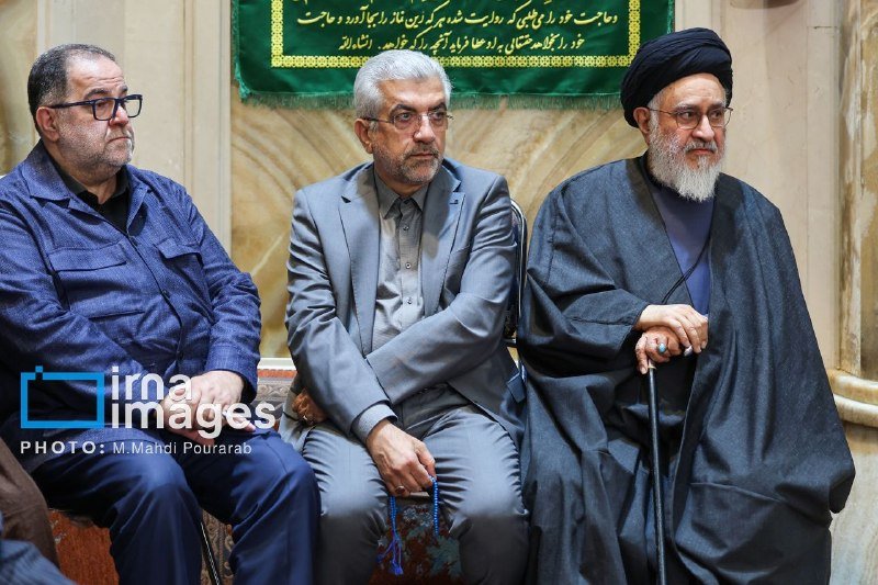 Three men sit side by side indoors, one holding prayer beads and another a cane, with a green Persian-script banner visible behind them.