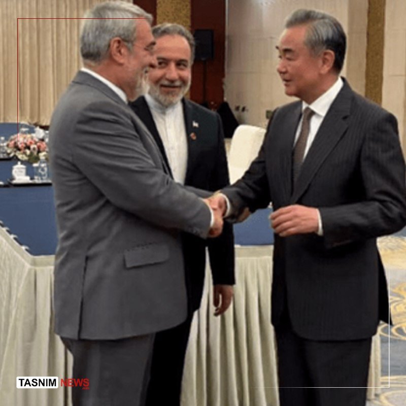 Three men in suits stand in a formal room, shaking hands and smiling near draped tables with floral arrangements.