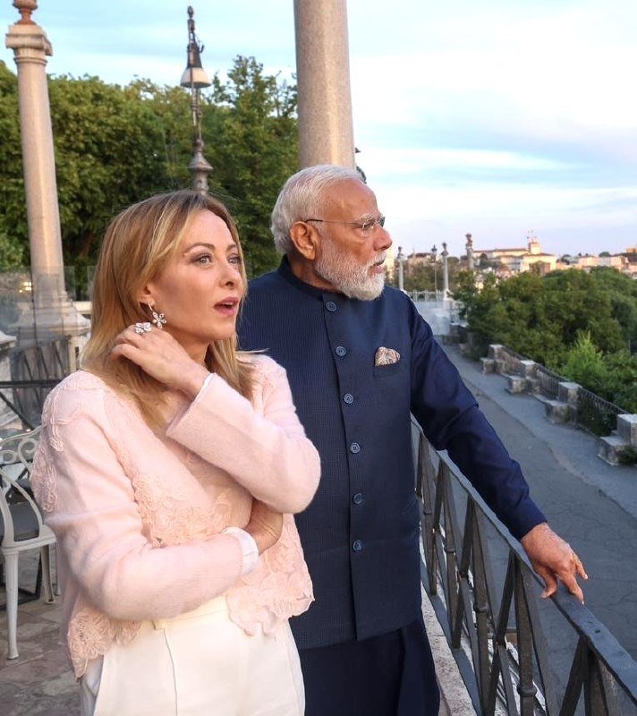 A blonde woman in a pink jacket and a white-bearded man in a dark blue vest stand on a balcony overlooking a tree-lined street with historic architecture.