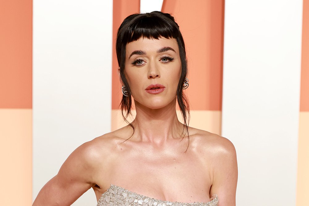 A woman with dark bangs and a strapless embellished gown poses against an orange and white backdrop.