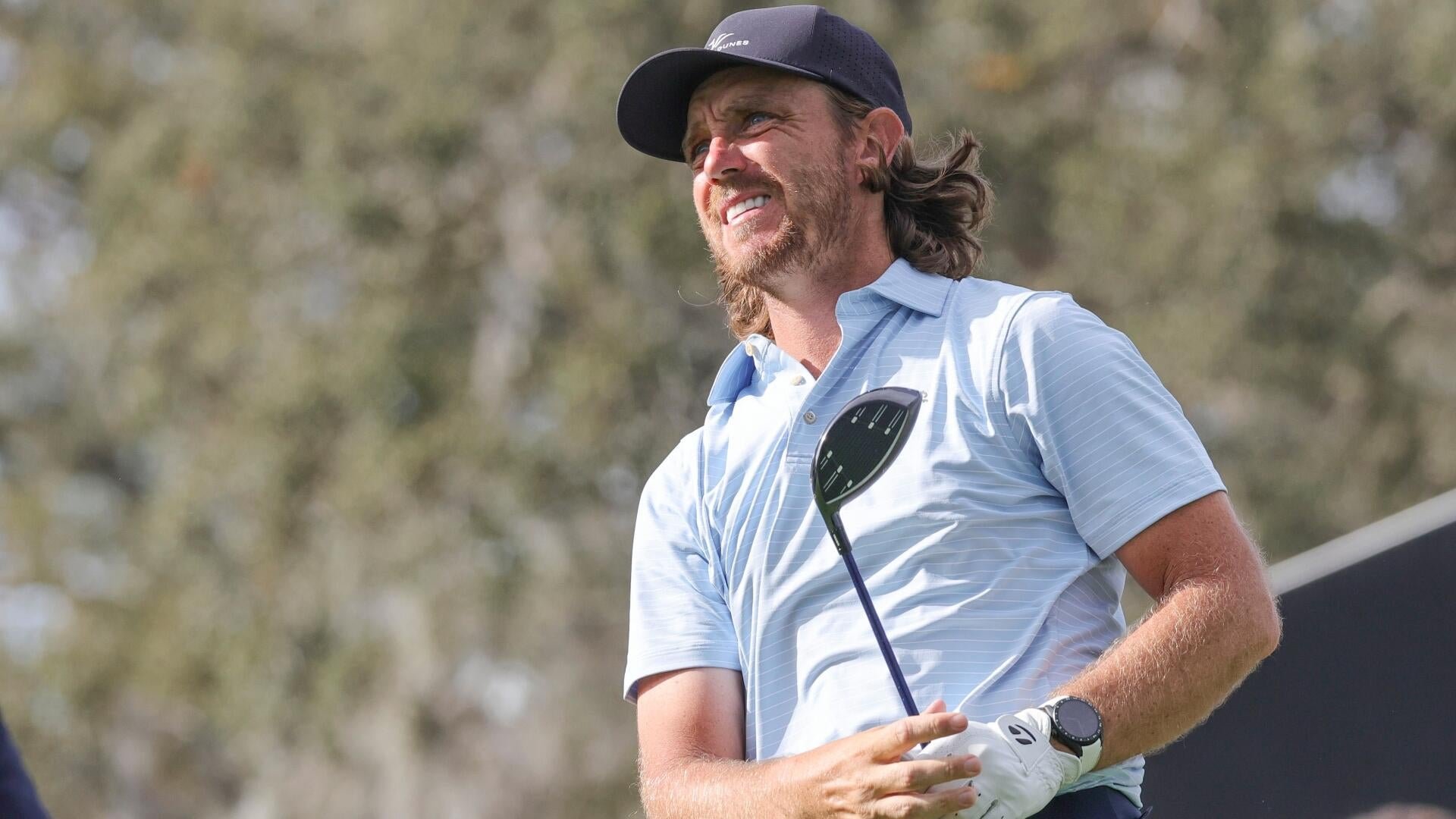 A bearded man wearing a navy cap and light blue striped polo shirt holds a golf club while looking upward with a smile against a backdrop of blurred trees.