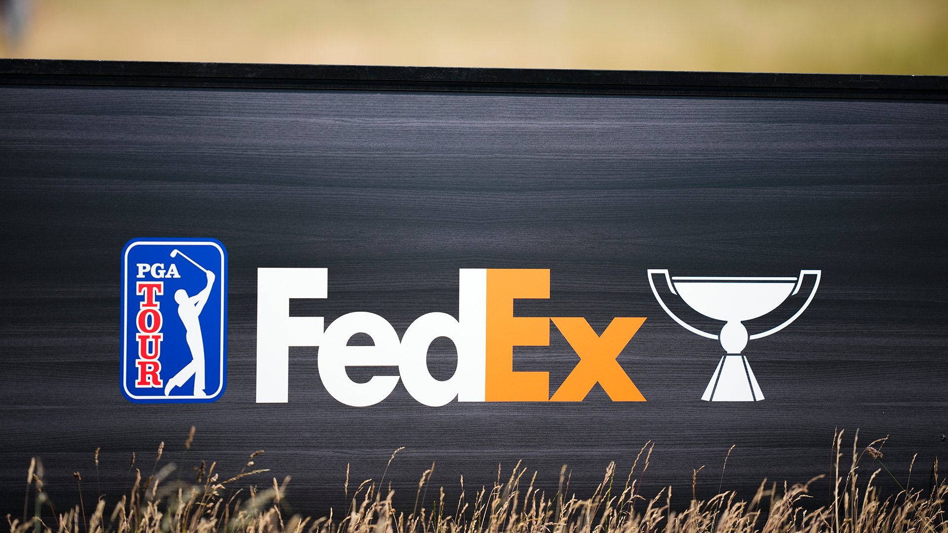 A black sign displays the PGA Tour logo, a FedEx wordmark, and a trophy icon, with tall grass visible in the foreground.