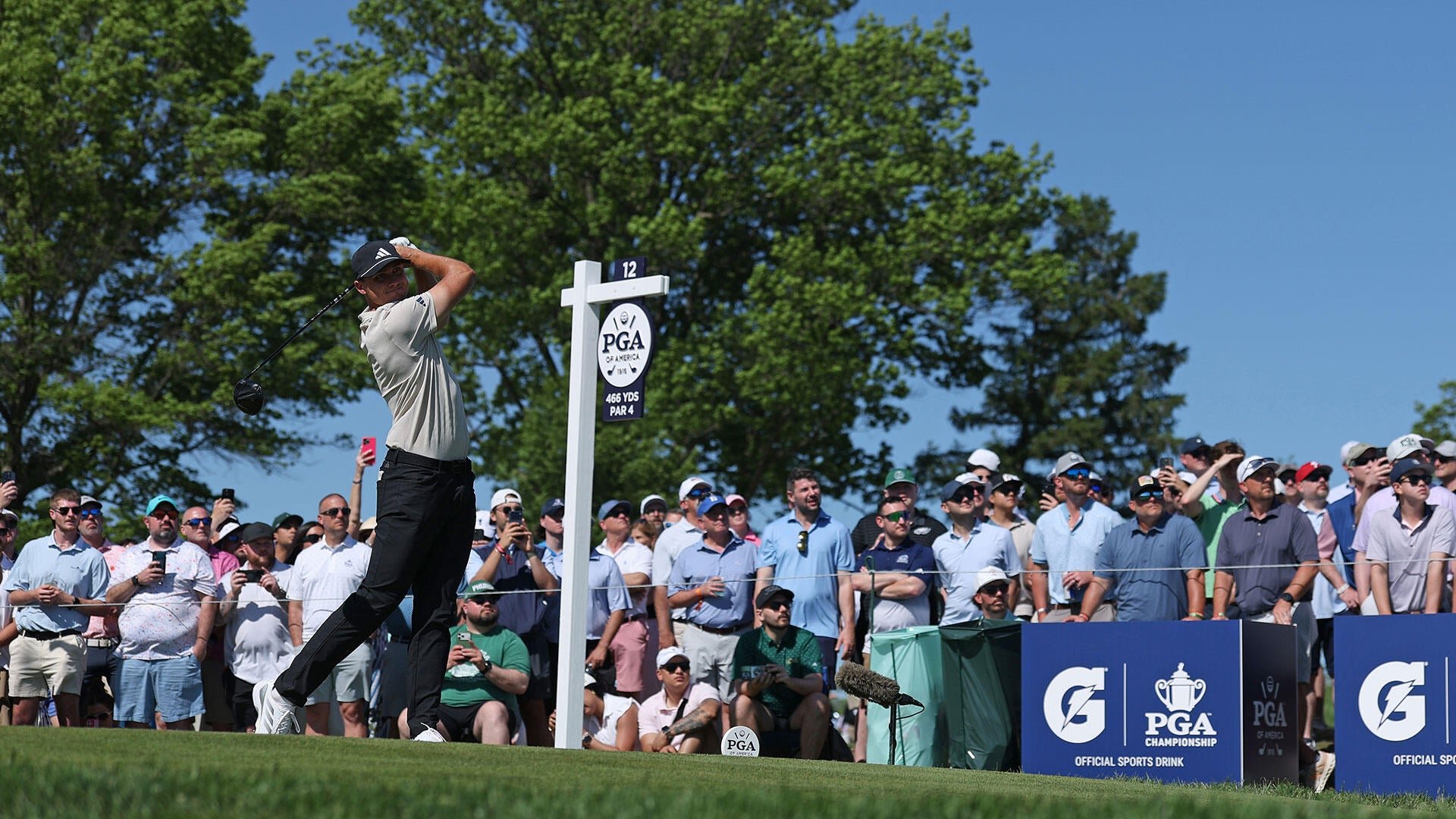 A golfer in a white shirt and black pants swings his club at the 12th tee (466 YDS, Par 4) while a crowd of spectators watches from behind a rope line.