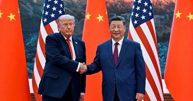 Two men in dark suits shake hands in front of alternating American and Chinese flags.
