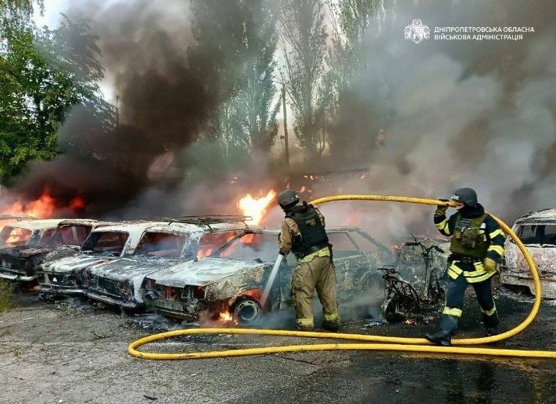 Two firefighters in helmets extinguish flames engulfing a row of burnt cars, with thick smoke rising and a yellow hose stretched across the scene.