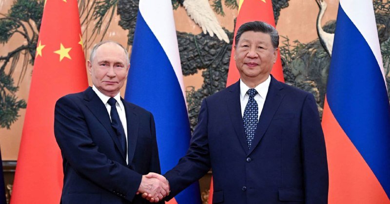 Two men in dark suits shake hands before alternating Chinese and Russian flags and an ornate decorative wall depicting cranes.
