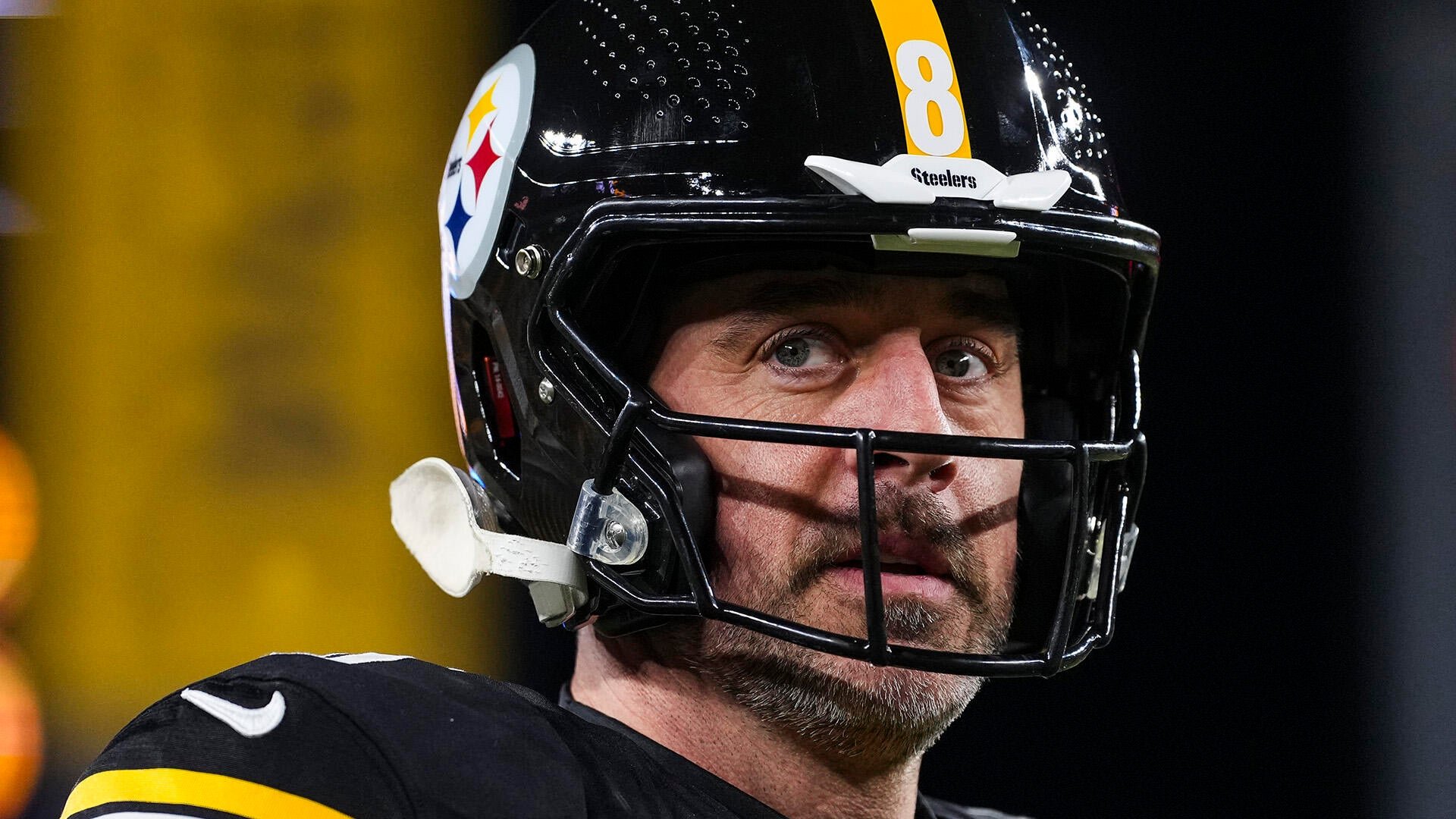 Close-up of a football player wearing a black Steelers helmet with a face mask, looking upward against a blurred yellow and dark background.