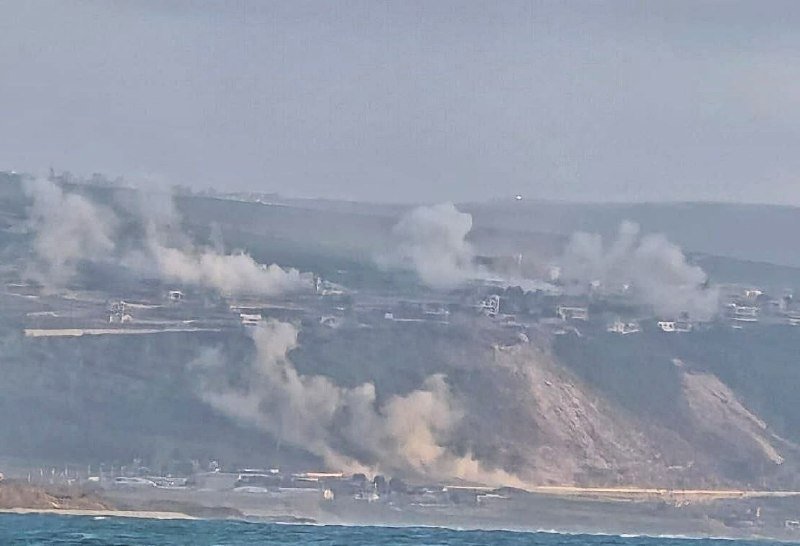 Plumes of smoke rise from multiple points across a coastal hillside with scattered buildings, viewed from across a body of water.