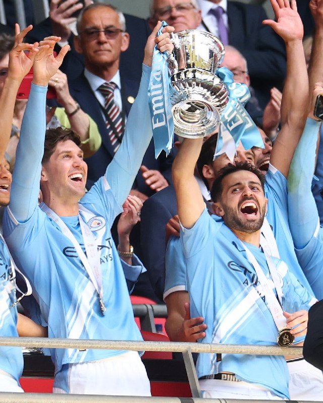 Two soccer players in light blue jerseys celebrate by hoisting a large silver trophy adorned with light blue ribbons reading "Emirates," wearing matching medals.