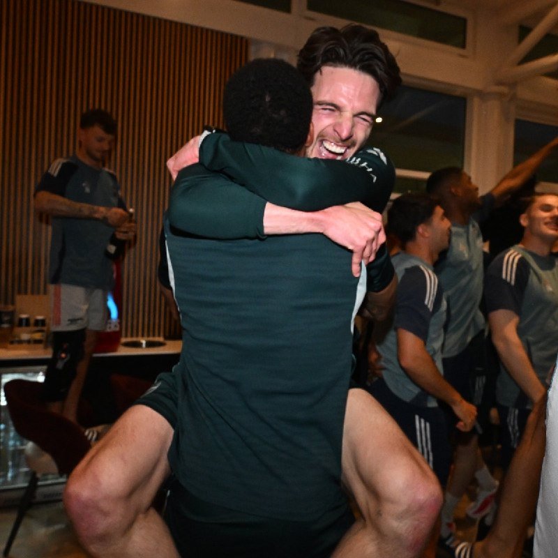 Two men in dark athletic shirts embrace and smile joyfully in a locker room while teammates in matching gear celebrate in the background.
