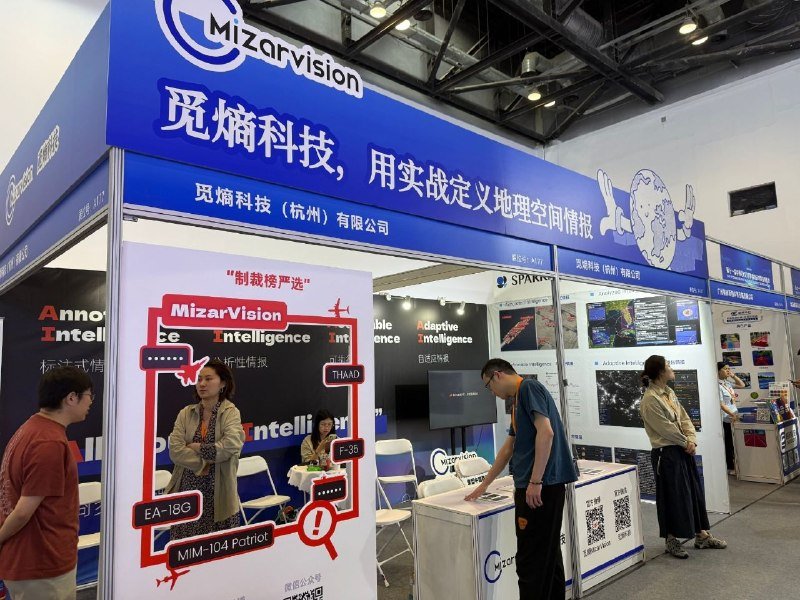 Attendees browse a blue MizarVision exhibition booth displaying digital screens, maps, and promotional graphics at a trade show.