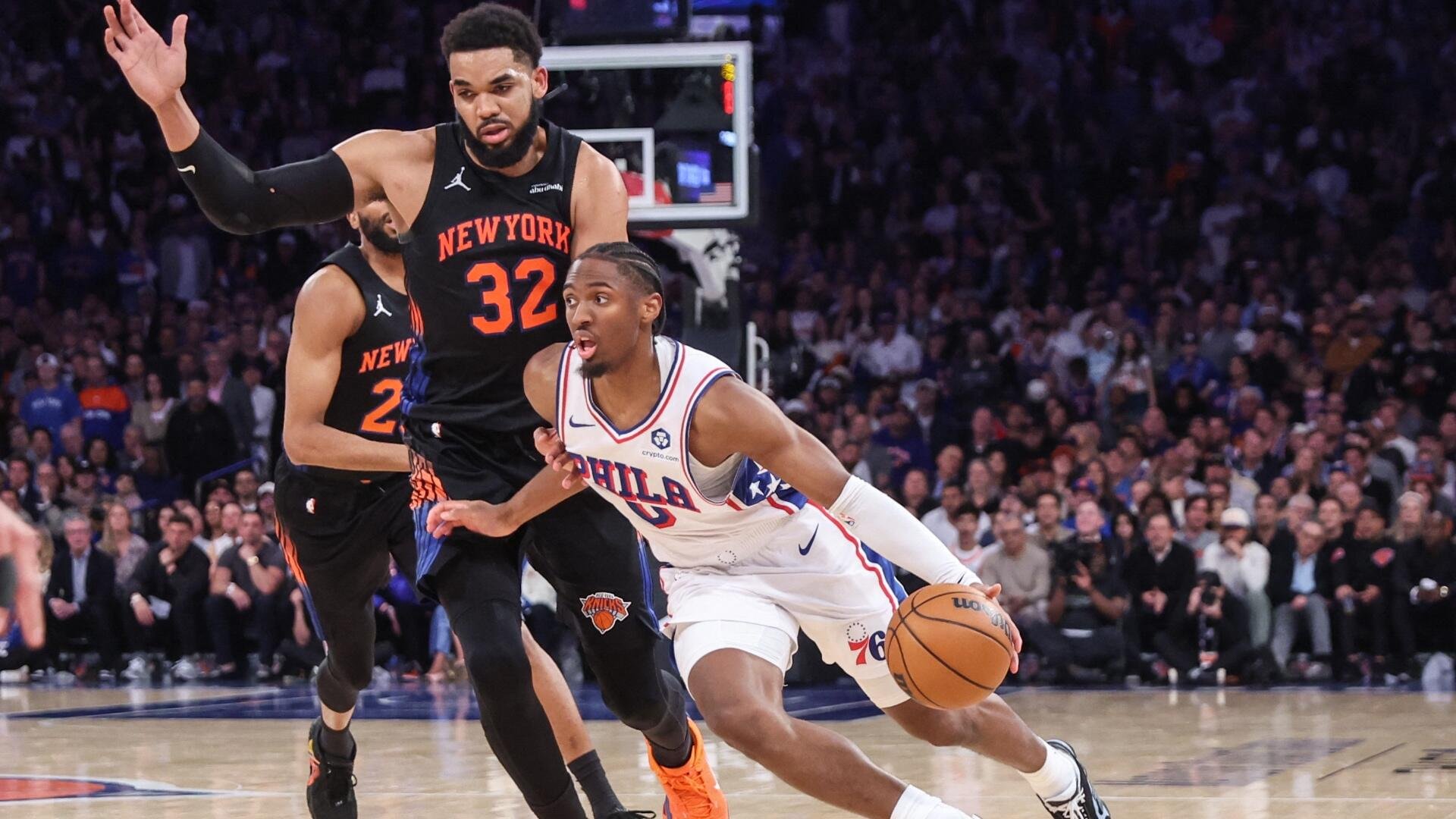A Philadelphia 76ers player in a white jersey dribbles past a New York Knicks defender wearing number 32 during an NBA game before a crowd.