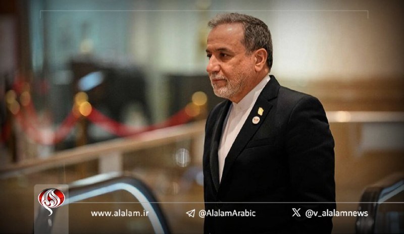 A man in a black suit stands in a hallway with an escalator behind him, with watermarks reading "www.alam.ir" and social media handles displayed.