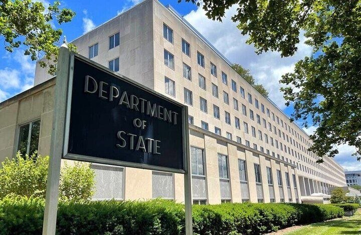 A large stone government building stands behind a black "Department of State" sign, surrounded by trees and hedges under a partly cloudy sky.
