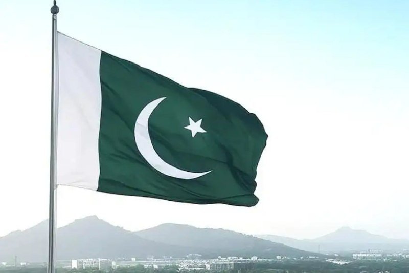 Pakistan's green and white flag with crescent and star waves on a flagpole against a hazy sky, with mountains and a cityscape visible in the background.