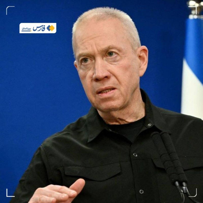 A gray-haired man in a black shirt speaks with a raised hand, a microphone visible, against a blue backdrop with a flag.