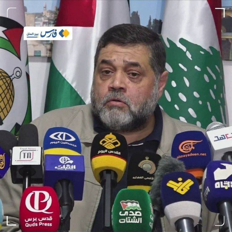 A bearded man speaks at a podium lined with numerous Arabic-labeled microphones, backed by flags including the Lebanese flag.
