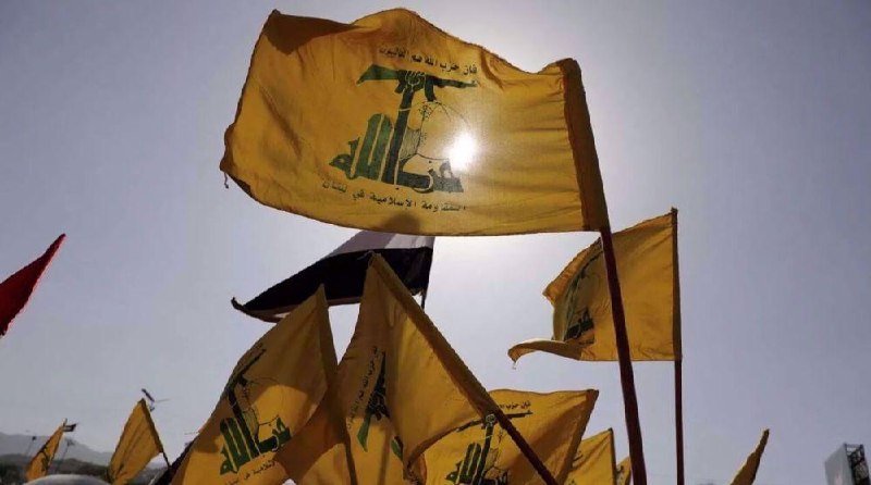 Multiple yellow flags with green Arabic script and an emblem flutter against a bright sky, with one red flag visible in the background.