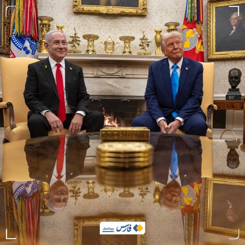 Two men in suits—one with a red tie, one with a blue tie—sit in yellow armchairs before a lit fireplace, with flags, gold ornaments, and framed portraits visible behind them.