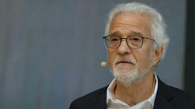 An older man with white hair, a beard, and glasses wears a headset microphone and speaks against a gray background.