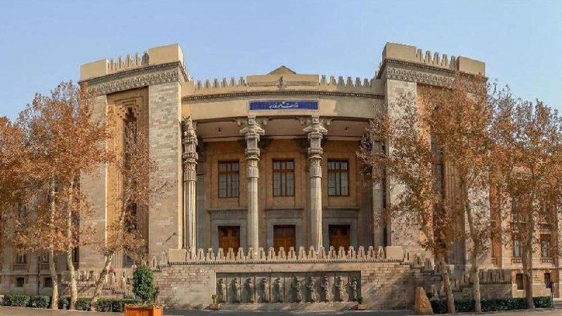A grand stone building featuring columns, crenellated towers, and Persian inscriptions stands flanked by autumn-colored trees under a clear sky.