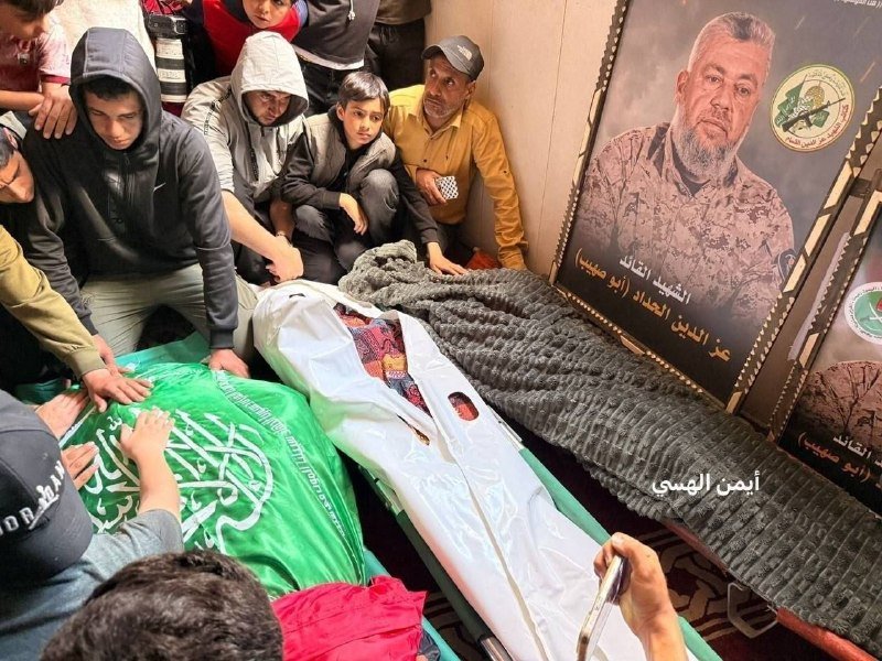 Mourners, including children and a man in a yellow shirt, gather around a body wrapped in a green Hamas flag and white shroud beside a framed portrait with Arabic text.