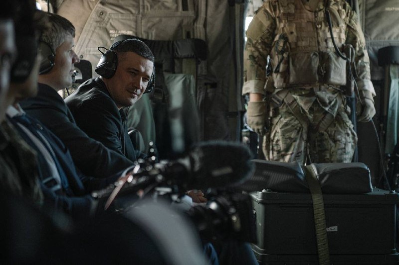 Inside a military aircraft, two men wearing headsets sit beside equipment cases while a third person in camouflage uniform stands nearby.