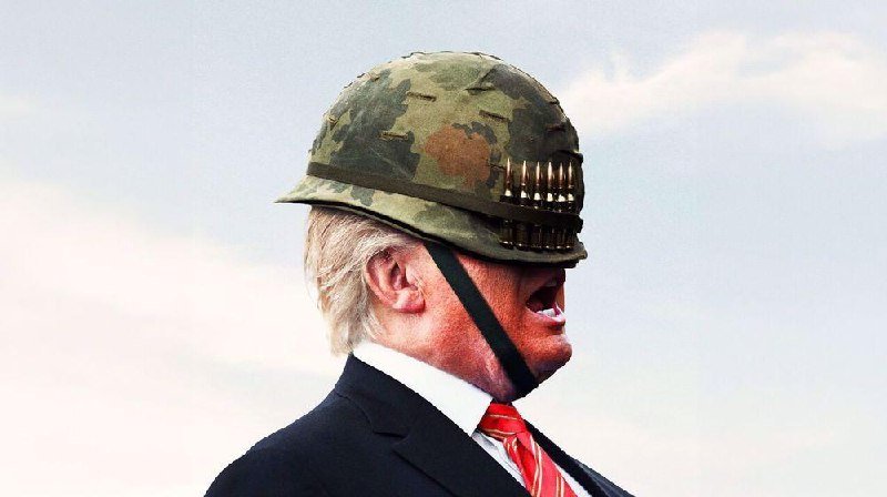 An older man with white hair, wearing a dark suit and red tie, speaks with his mouth open while wearing a camouflage military helmet against a cloudy sky.