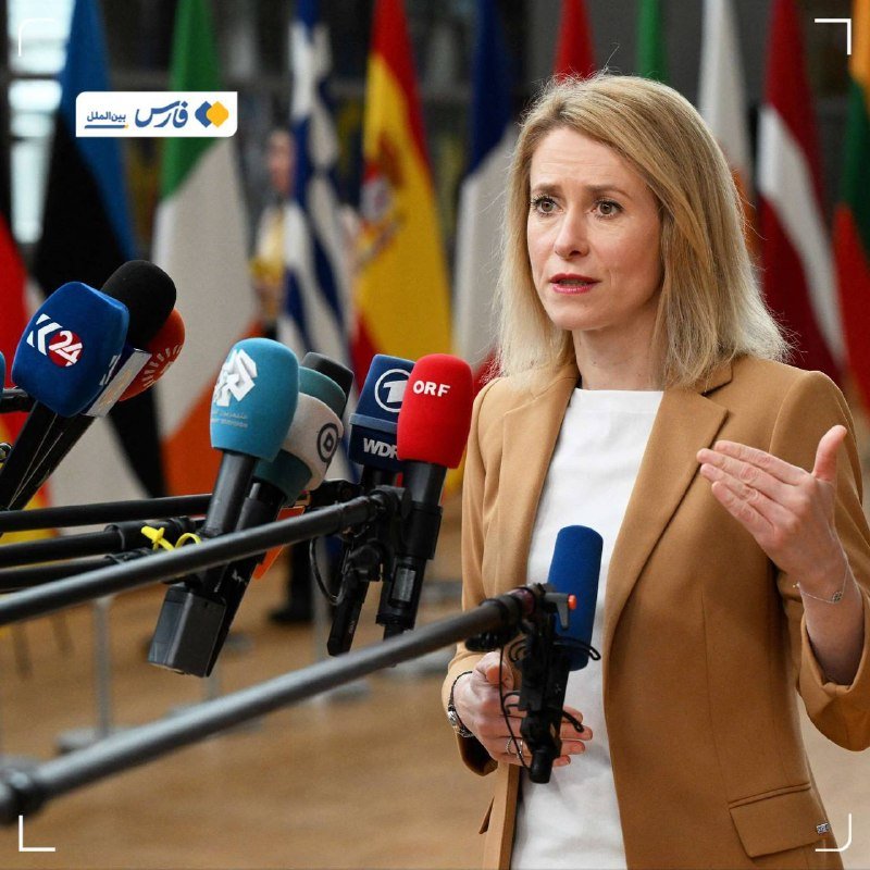 A blonde woman in a tan blazer speaks to multiple microphones labeled with various news outlets, gesturing with her hand, against a backdrop of international flags.