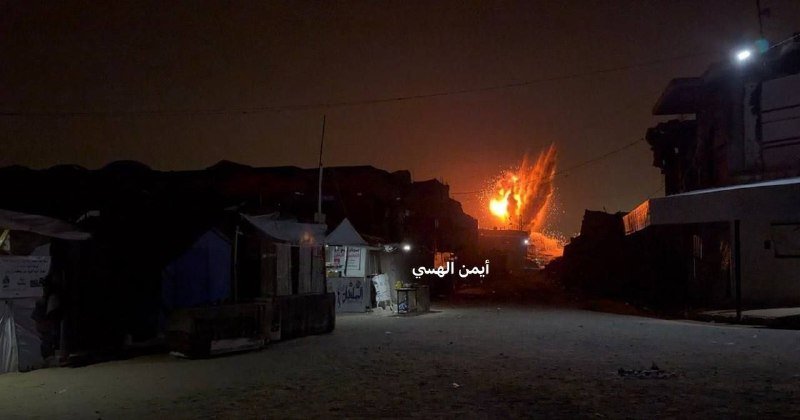 A nighttime scene shows a large fire burning in the distance, illuminating the dark sky above a row of dimly lit buildings and an empty street.