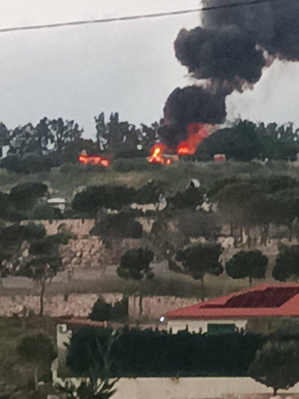 Thick black smoke rises from a fire burning on a hillside dotted with trees, above a house with a red tile roof in the foreground.