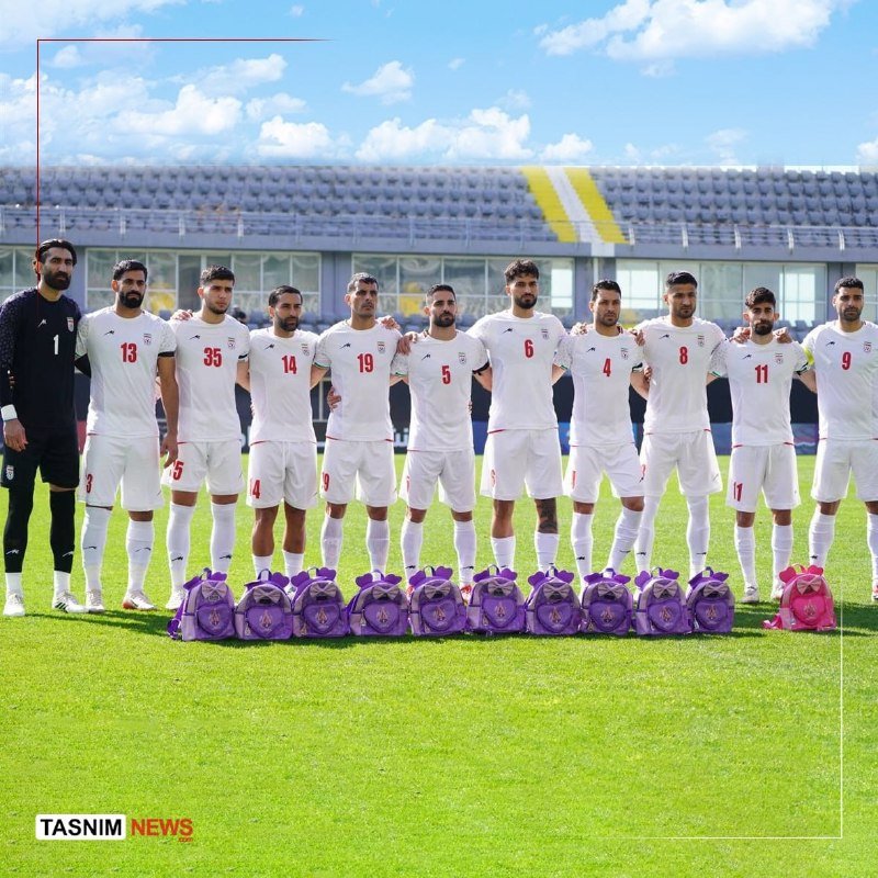 A soccer team in white uniforms lines up on a grass field with a row of purple bags placed in front of them, one goalkeeper in black standing at the end.
