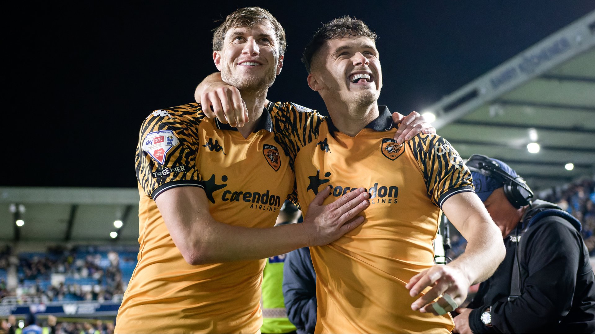 Two smiling soccer players in orange and black striped Hull City jerseys embrace in celebration on a stadium pitch at night.
