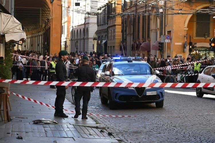 Two uniformed officers and a police car with flashing lights stand behind red-and-white tape, blocking a crowd from a fish lying on the cobblestone street.