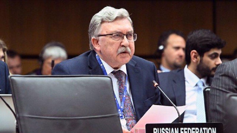 A gray-haired man in a suit and glasses sits at a desk labeled "RUSSIAN FEDERATION," speaking into a microphone during a formal conference setting.