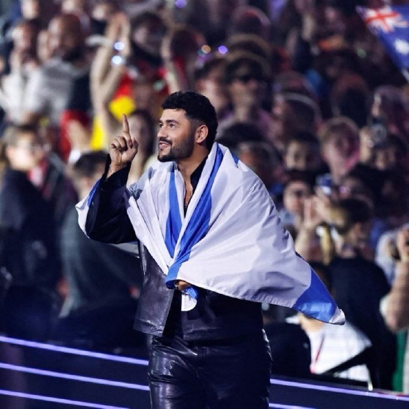 A man wearing a white and blue flag draped over his shoulders points upward while standing before a large crowd holding cameras.
