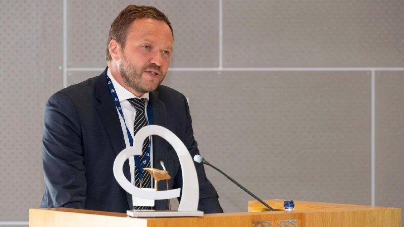 A bearded man in a dark suit and striped tie speaks at a wooden podium beside a sculptural award and a microphone.