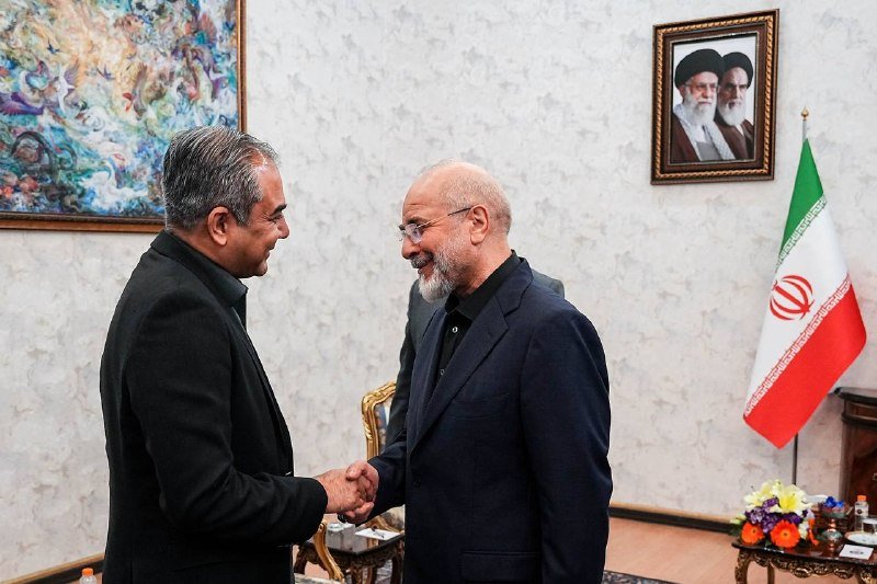Two men in dark suits shake hands and smile in a formal room featuring an Iranian flag and a framed portrait of two clerics on the wall.