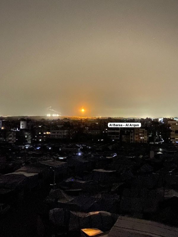A bright orange flash illuminates the night sky over a darkened urban landscape, with city lights glowing in the distance.