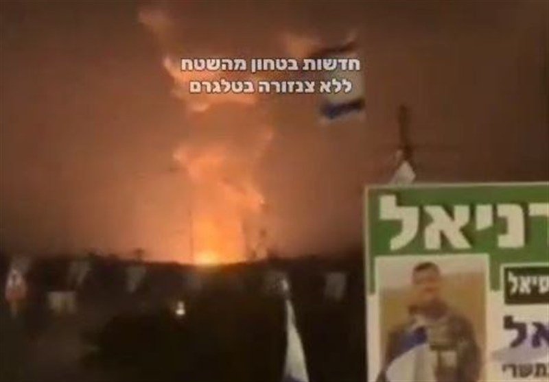 Large flames and thick smoke rise from a fire at night, with an Israeli flag and Hebrew political campaign signs visible in the foreground.