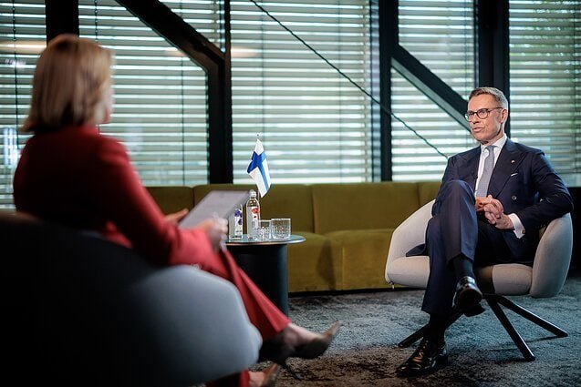 A man in a suit and glasses sits across from a woman in a red jacket during an interview, with a small Finnish flag on the table between them.