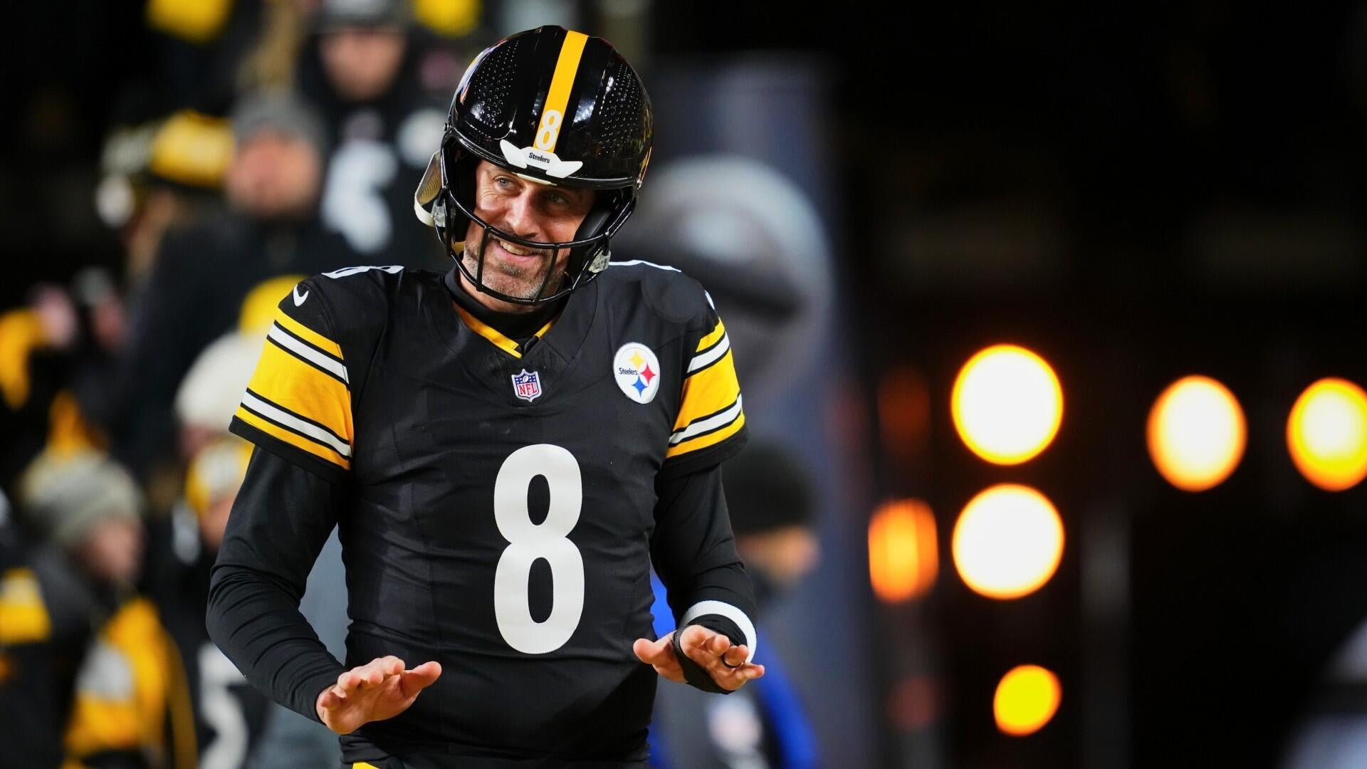 A football player wearing a black jersey with the number 8 and a Steelers helmet smiles while gesturing with both hands, with blurred fans and stadium lights behind him.