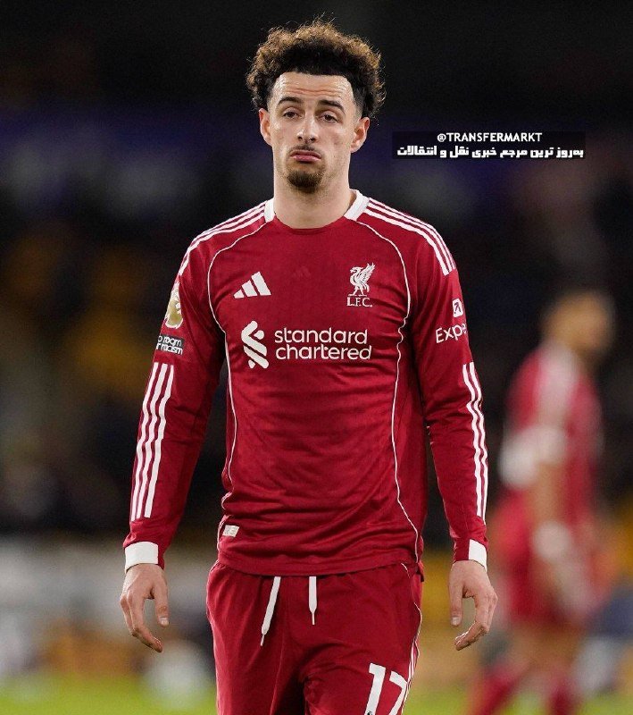 A young player with curly hair walks forward in a red Liverpool FC jersey with "Standard Chartered" sponsorship, on a soccer pitch.