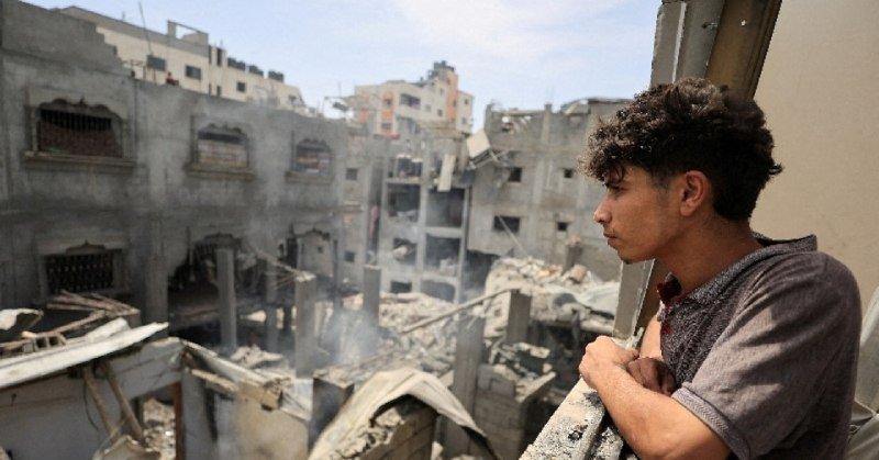 A young man stands on a balcony looking out over a landscape of heavily damaged and collapsed buildings with smoke rising from the rubble.