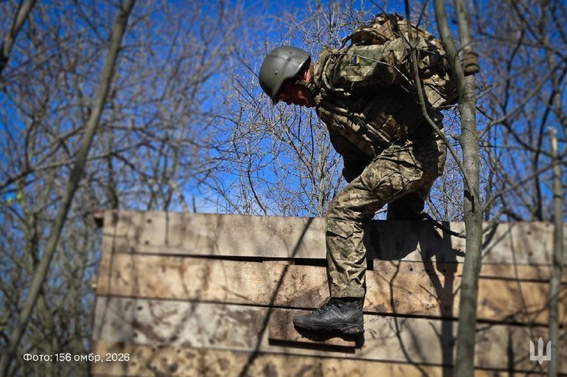 A soldier in camouflage uniform and helmet climbs over a wooden barrier outdoors, carrying a backpack, with a small Ukrainian flag patch visible on the uniform.