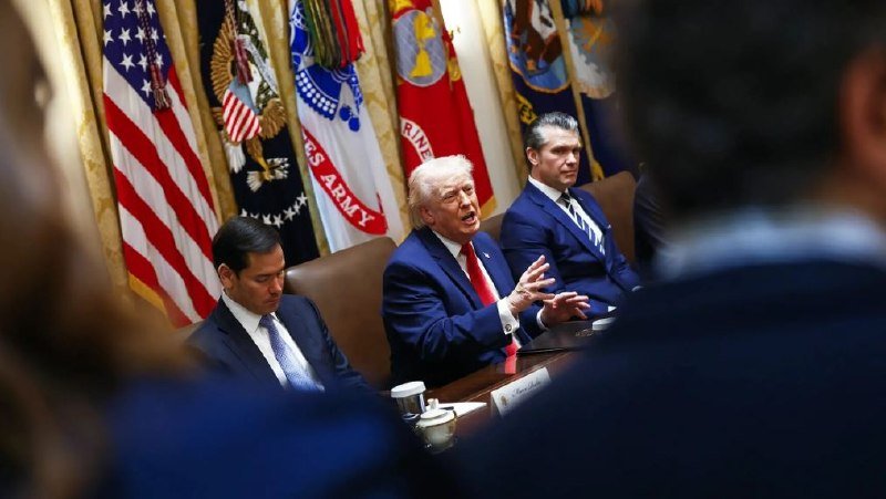 Three men in suits sit at a table in front of U.S. and military flags, with the center man gesturing while speaking.