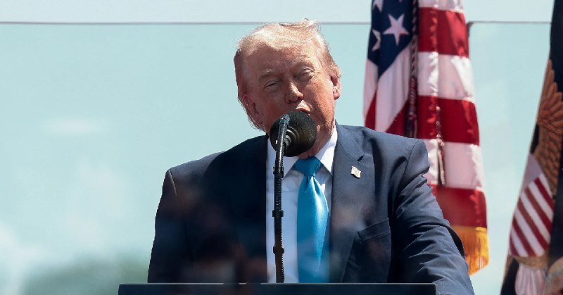 A man with blonde hair in a dark suit and blue tie speaks at a podium with a microphone, with an American flag visible in the background.