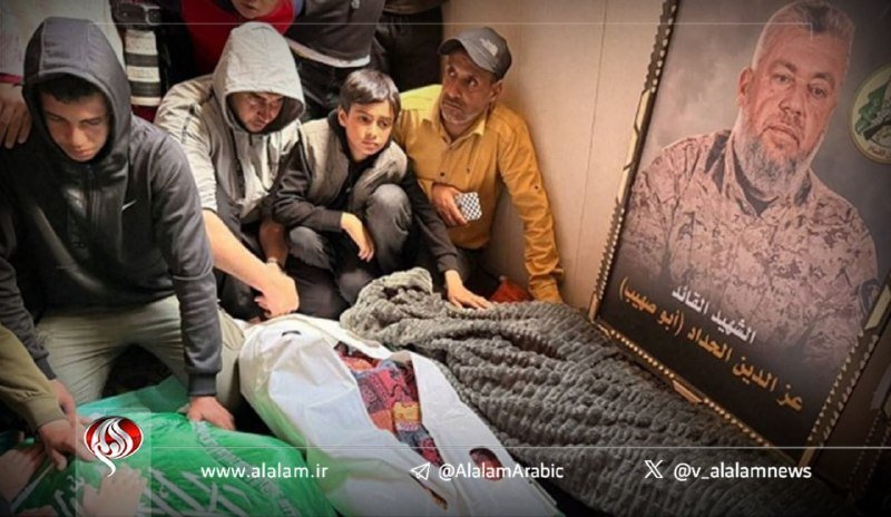 Mourners, including children and young men, gather around a body wrapped in a green and white cloth beside a framed portrait of a man with Arabic text.