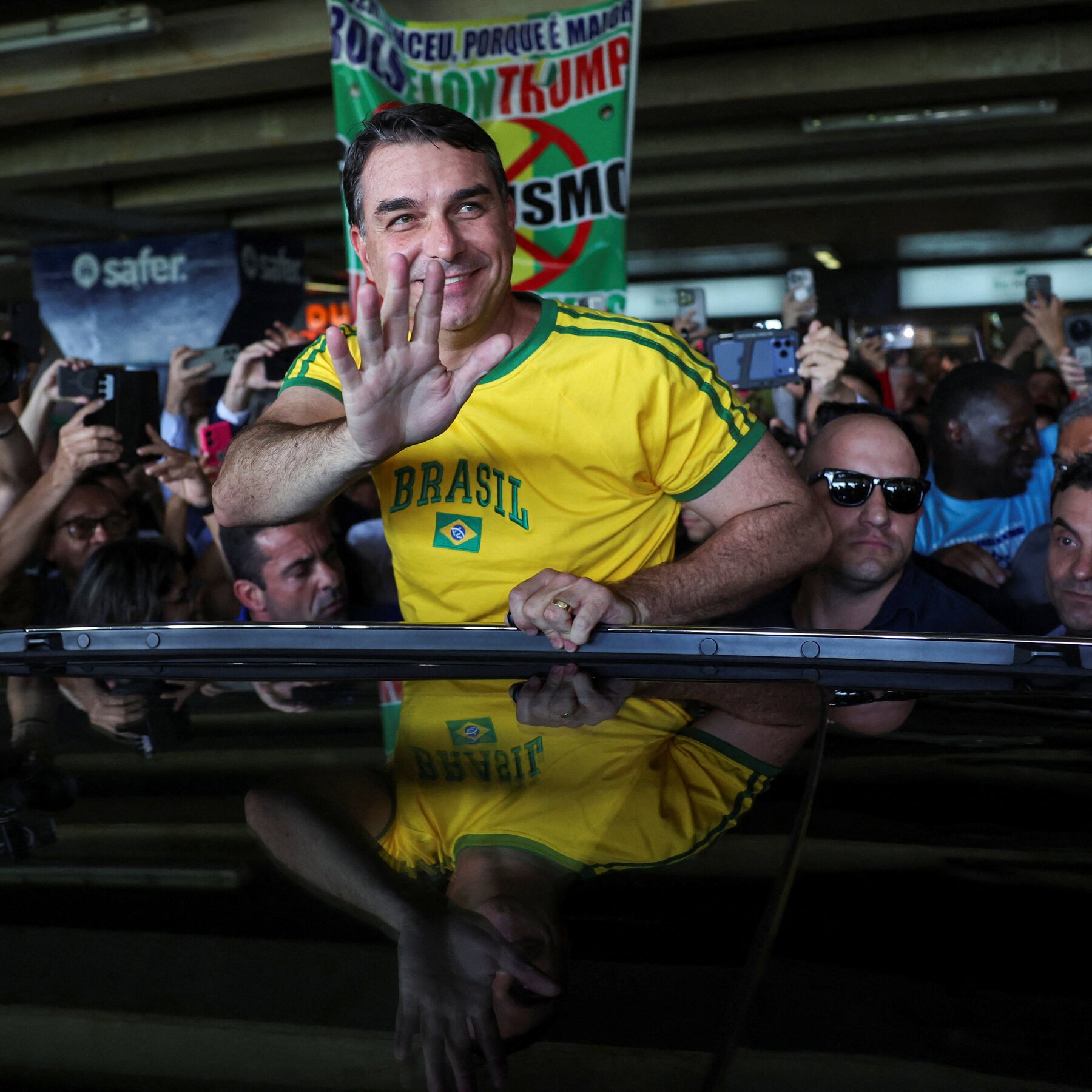 A smiling man in a yellow "Brasil" jersey waves beside a black vehicle, surrounded by a crowd holding up phones to photograph him.
