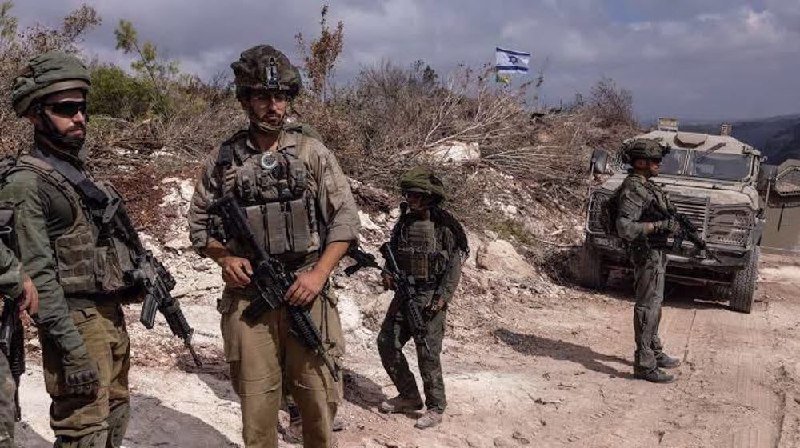 Four armed soldiers in combat gear stand near a military vehicle on a dirt road, with an Israeli flag visible in the background.
